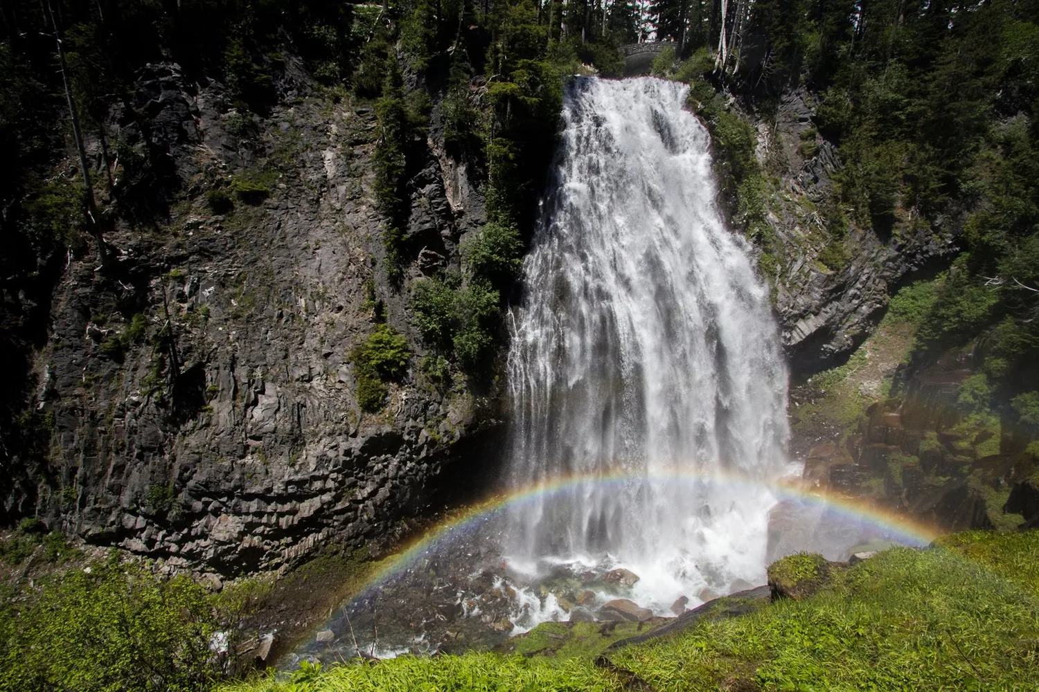 A fan-shaped waterfall cascades over lava cliffs with a rainbow in its mist.