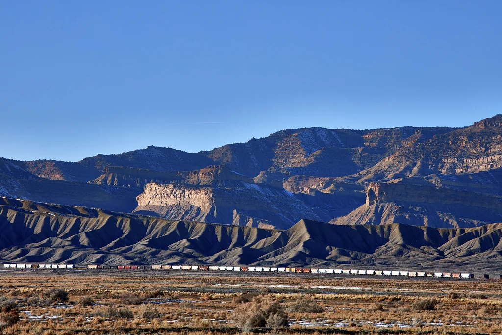 A classic long freight train (many of which exceed 100 cars) cross a remote, mostly dry valley near the small city of Green River (population c. 950 as of 2022) in the western U.S. state of Utah